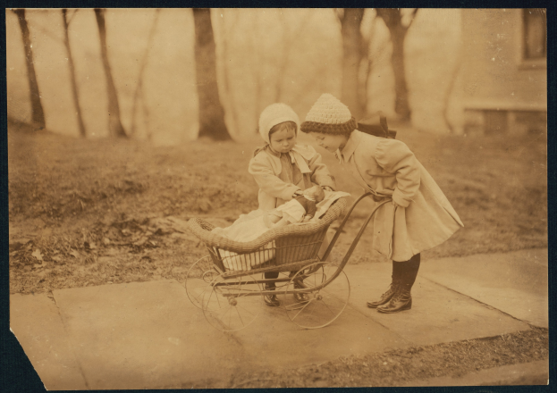 Zwei Kinder in Mänteln und Mützen schieben ein Baby in einem Kinderwagen über eine grüne Fläche mit Bäumen und einem Gebäude im Hintergrund.