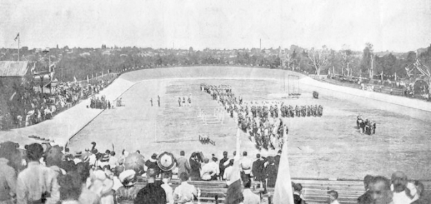 Ein altes Schwarz-Weiß-Foto einer Menge, die ein Pferderennen in einem Stadion betrachtet, mit Menschen auf Bänken sitzend, stehend, Fahnen haltend und reitend, mit Bäumen und einem klaren Himmel im Hintergrund.
