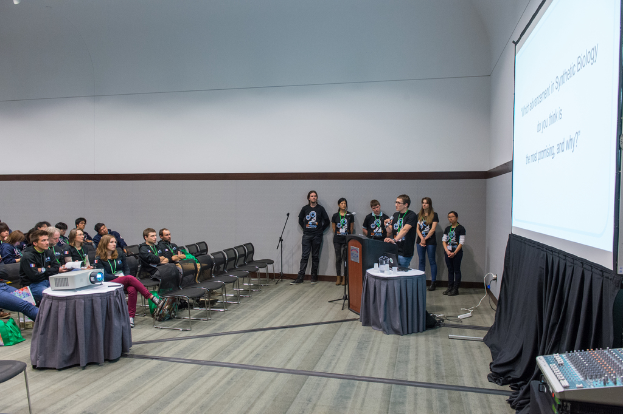 Eine Person spricht in ein Mikrofon in einem Bürosetting, mit Zuschauern und Teammitgliedern mit ID-Karten dahinter, einer Präsentation und einem Videoplayer.