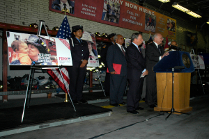 Eine Gruppe von Menschen steht in der Nähe eines Podiums mit Mikrofonen, Schildern auf Ständern, Fahnen und Schildern an der Wand, alles unter hellem Licht.