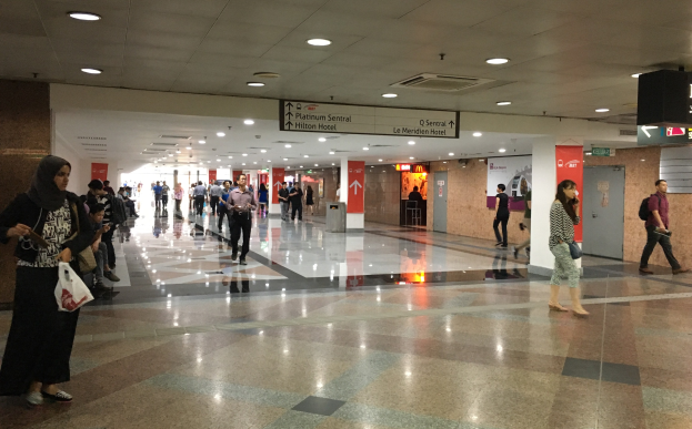 Menschen mit Taschen durch eine Flughafenhalle gehend, mit Informationsschildern an den Wänden, Deckenleuchten und einem Mülleimer im Hintergrund; eine Frau ist in Schuhen zu sehen.