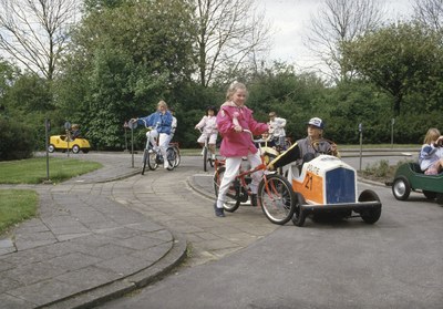 Eine Gruppe von Kindern, die Fahrräder auf einer von Bäumen gesäumten Straße hinunterfahren, mit einer Person auf einem Fahrrad und einem Auto im Vordergrund, einem klaren blauen Himmel im Hintergrund.