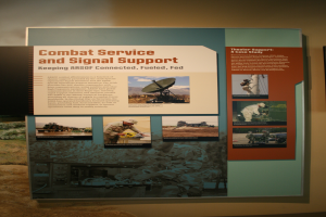 Ein buntes Plakat mit einem blauen Hintergrund und weißer Schrift, das Bilder von Militärpersonal in Aktion zeigt und den Text "Kampfunterstützung und Signalsupport" in fetter, schwarzer Schrift enthält.