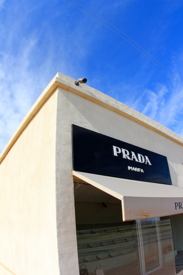 Ein Namensschild an einer Gebäudewand mit einer Sicherheitskamera und Glastüren, mit einem blauen Himmel und Wolken im Hintergrund.