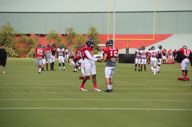 Gruppe von Menschen, die auf einem Rasenfeld Fußball spielen, mit Helmen und Stiefeln, Bäumen und Strukturen im Hintergrund unter einem klaren blauen Himmel.