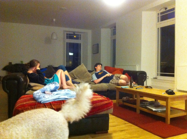 Ein Wohnzimmer mit einer Couch, einem Tisch, T├╝ren und Fenstern, auf dem eine Hündin, ein Mann und eine Frau sitzen, sowie diverse Gegenst├Ąnde wie eine Wasserflasche, eine Fernbedienung, B├╝cher, Papiere und Stifte.