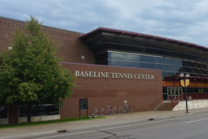 Das Baseline Tennis Center in St. Louis, Missouri, ein großes Gebäude mit Glasfenstern und einem Namensschild, mit Treppen und Geländern, die zum Eingang führen, Fahrräder vor dem Gebäude geparkt, ein Baum links, eine Straße unten und ein bewölkter Himmel im Hintergrund.