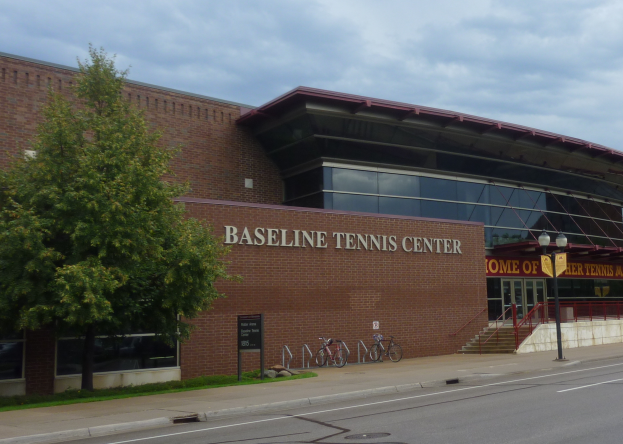 Das Baseline Tennis Center in St. Louis, Missouri, ein großes Gebäude mit Glasfenstern und einem Namensschild, mit Treppen und Geländern, die zum Eingang führen, Fahrräder vor dem Gebäude geparkt, ein Baum links, eine Straße unten und ein bewölkter Himmel im Hintergrund.