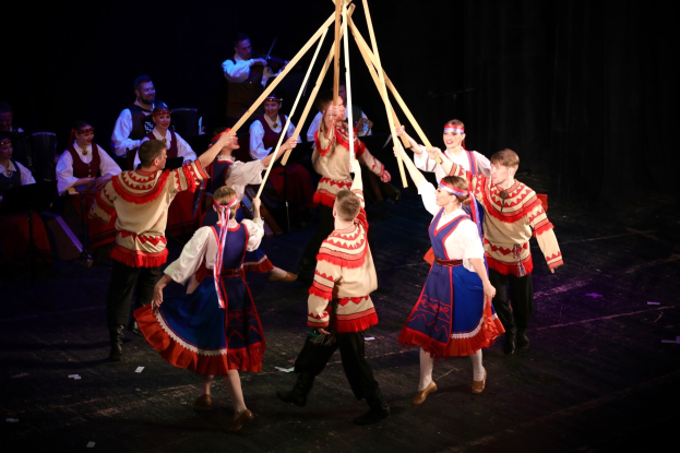 Eine Gruppe von Menschen in farbenfrohen Kostümen tanzt um einen Maibaum, einige halten Holzstäbe, während andere Musikinstrumente im Hintergrund spielen.