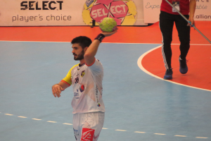 Ein Mann hält einen Handball auf einem Feld, neben ihm steht eine Person mit einem Stock, im Hintergrund sind Banner mit Text und Logos.