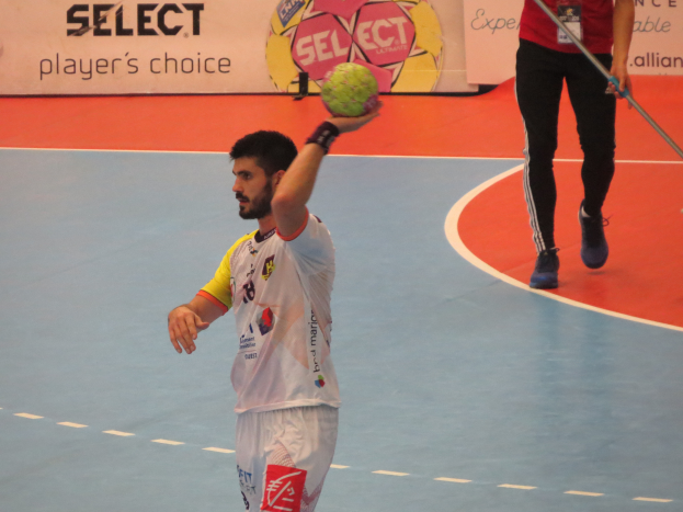 Ein Mann hält einen Handball auf einem Feld, neben ihm steht eine Person mit einem Stock, im Hintergrund sind Banner mit Text und Logos.