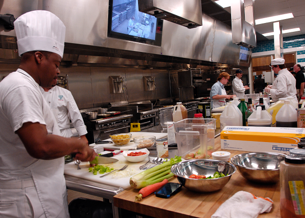 Eine Gruppe von Köchen in einer Küche, mit einem in Weiß gekleidetem Koch im Vordergrund, der ein Messer hält und von verschiedenen Utensilien, Schüsseln, Gemüse, einem Handy, Flaschen und anderen Gegenständen umgeben ist, mit einem Fernseher und Deckenleuchten im Hintergrund.