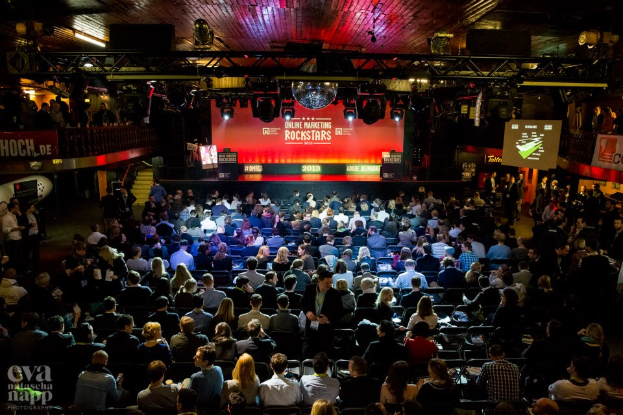 Eine große Menschenmenge, die auf Stühlen vor einer Bühne mit Bildschirmen und Bannern sitzt, die von Scheinwerfern beleuchtet wird und von Geländern umgeben ist, erwartet gespannt den Beginn eines Online-Marketing-Rockstars-Events, mit einem Wasserzeichen in der linken unteren Ecke.