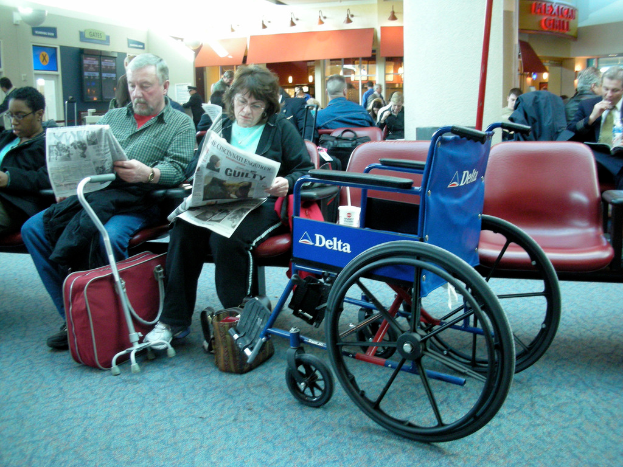 Mehrere Personen sitzen auf Stühlen, mit einem Rollstuhl im Vordergrund und zwei Individuen mit Zeitungen; ein mittlerer Balken mit Lampen ist in der Mitte sichtbar.