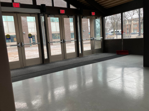 Ein großer Raum mit vielen Fenstern und Türen, ein Besen auf dem Boden, Schilder, ein Dach, Bäume, Fahrzeuge, Gebäude und ein bewölkter Himmel, der eine kommerzielle Bodenreinigungsdienstleistung darstellt.