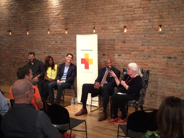 Eine Gruppe von Menschen in eleganter Kleidung sitzt auf Stühlen in einem Raum mit einer roten Backsteinwand und Plakaten im Hintergrund und lauscht einer Frau rechts, die in ein Mikrofon singt.