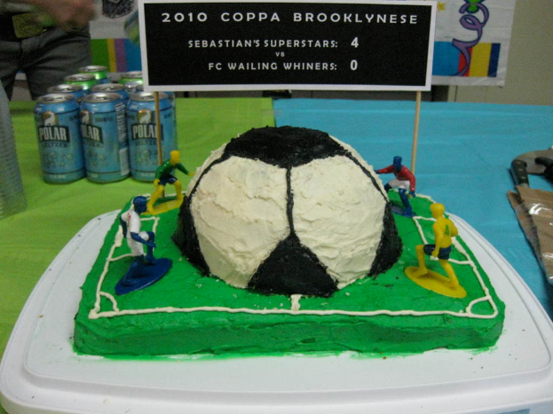 Ein Fußballstadion-förmiger Kuchen mit einem Fußball und vier Spielern, umgeben von Softdrink-Dosen, mit einem Banner im Hintergrund und einer Person auf der oberen linken Seite, mit einer Platte im Kuchen.