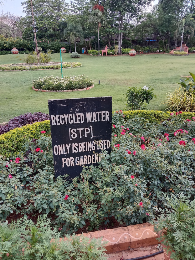 Ein Garten mit einem Schild, auf dem "Recycled Water (STP) Only Being Used for Gardening" steht, umgeben von grünem Gras, Blumen, Bäumen und einem blauen Himmel.