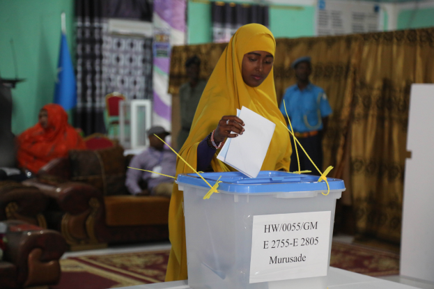 Eine Frau mit gelbem Kopftuch gibt ihre Stimme in einer Wahllokal ab, vor einem Tisch mit einer Box stehend, während einige Leute auf der linken Seite sitzen, mit Vorhängen, einer Flagge und einer Wand im Hintergrund.