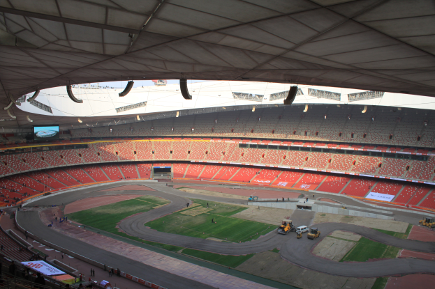 Ein großes Stadion voller Menschen, Fahrzeuge auf der Straße, Gras auf dem Boden, Schilder mit Text, Geländer, ein Bildschirm und eine Decke mit Lampen oben.