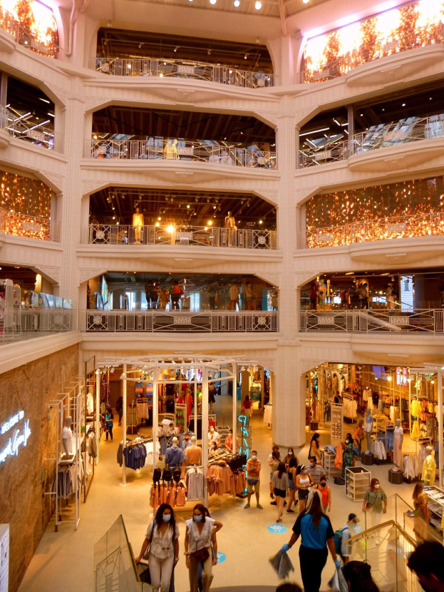 Großer Atrium eines belebten Einkaufszentrums mit Menschen, die Taschen tragen und Schaufensterpuppen mit Kleidern anschauen, umgeben von Geländern, Beleuchtung und Wandtafeln.