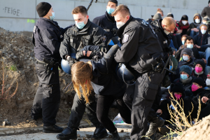 Gruppe maskierter Individuen in Käpfen, einige mit Waffen, die eine Frau umgeben, die von der Polizei eskortiert wird, mit Pflanzen, Steinen und einer Wand mit Protesttext im Hintergrund.