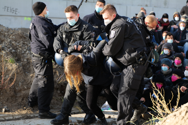 Gruppe maskierter Individuen in Käpfen, einige mit Waffen, die eine Frau umgeben, die von der Polizei eskortiert wird, mit Pflanzen, Steinen und einer Wand mit Protesttext im Hintergrund.