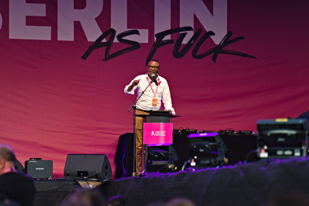 Ein Mann in einem weißen Hemd und braunen Hosen steht auf einer Bühne und hält ein Mikrofon in der Hand, mit einem pinken 'Berlin as Fuck'-Schild hinter ihm und einer Menge vor ihm.