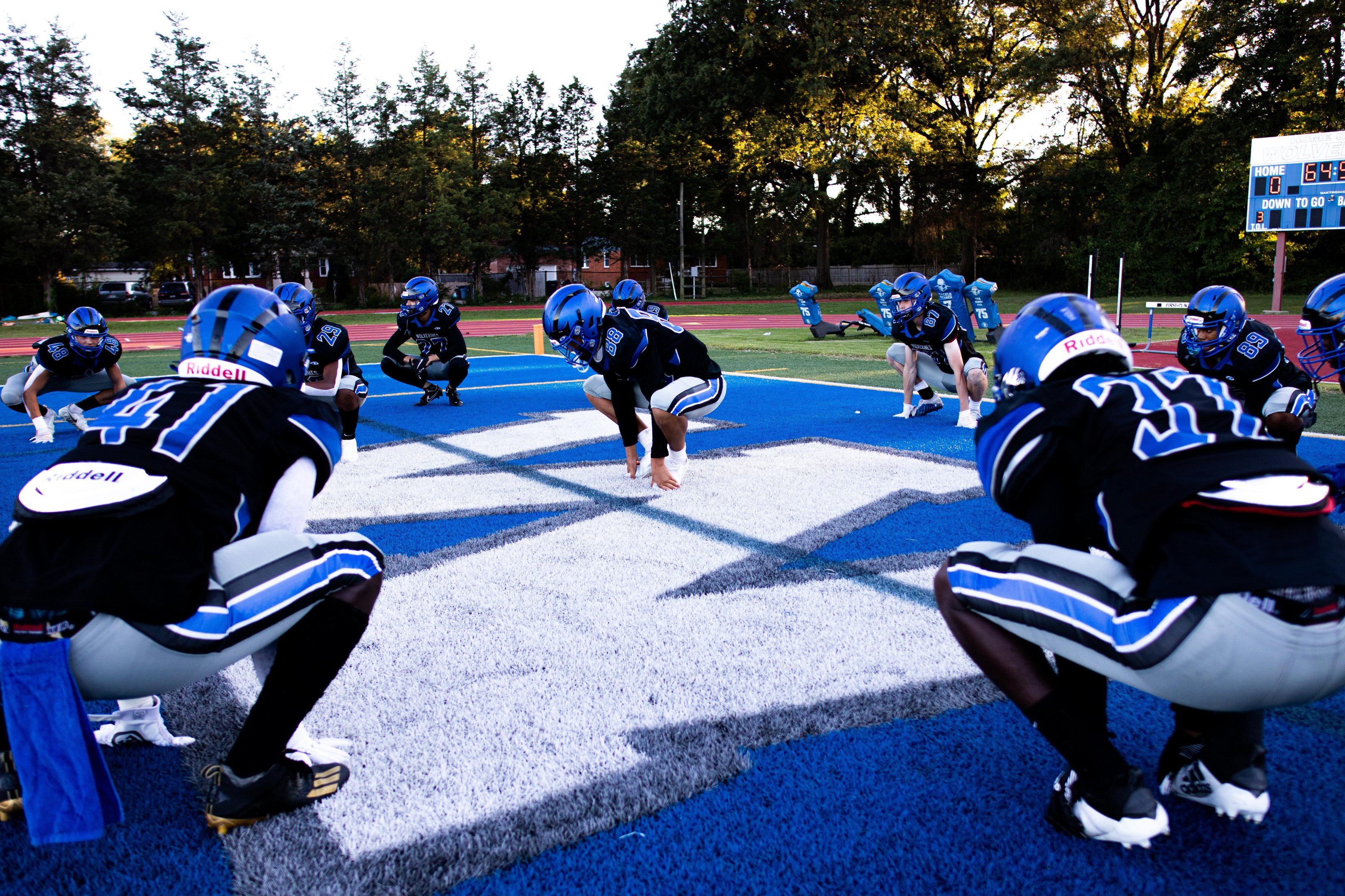 Junge Männer in Sportkleidung und Helmen beim Football-Spiel auf einem Feld mit einem Scoreboard und Bäumen im Hintergrund unter einem klaren blauen Himmel.