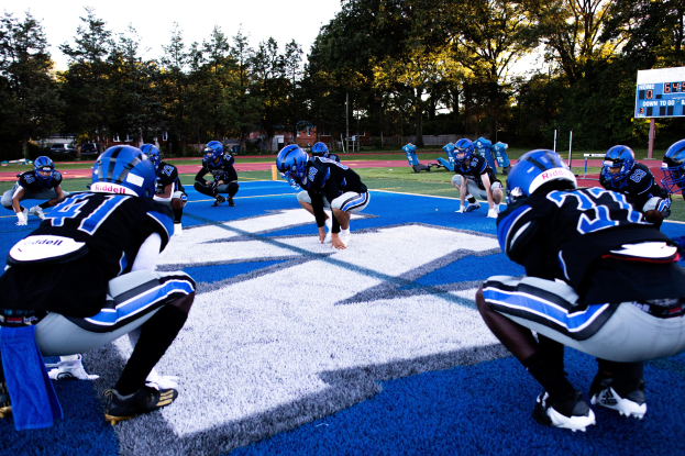 Junge Männer in Sportkleidung und Helmen beim Football-Spiel auf einem Feld mit einem Scoreboard und Bäumen im Hintergrund unter einem klaren blauen Himmel.