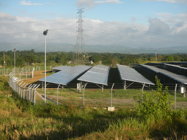 Ein Solarpanelfeld, das von einem Zaun umgeben ist, mit Gras, Pflanzen und Bäumen im Vordergrund, vor einem wolkenverhangenen Himmel und einem Übertragungsturm mit Drähten im Hintergrund.