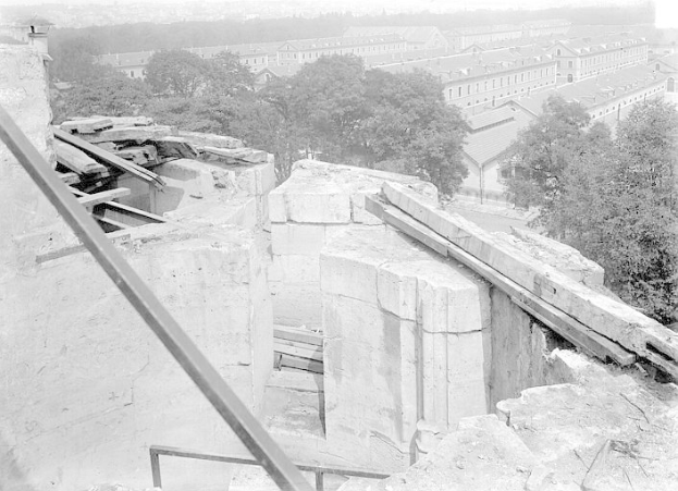 Ein Schwarz-Wei├č-Foto eines teilweise abgerissenen Geb├Ąudes mit verstreuter Tr├╝mmern, B├Ąumen und anderen Geb├Ąuden im Hintergrund.
