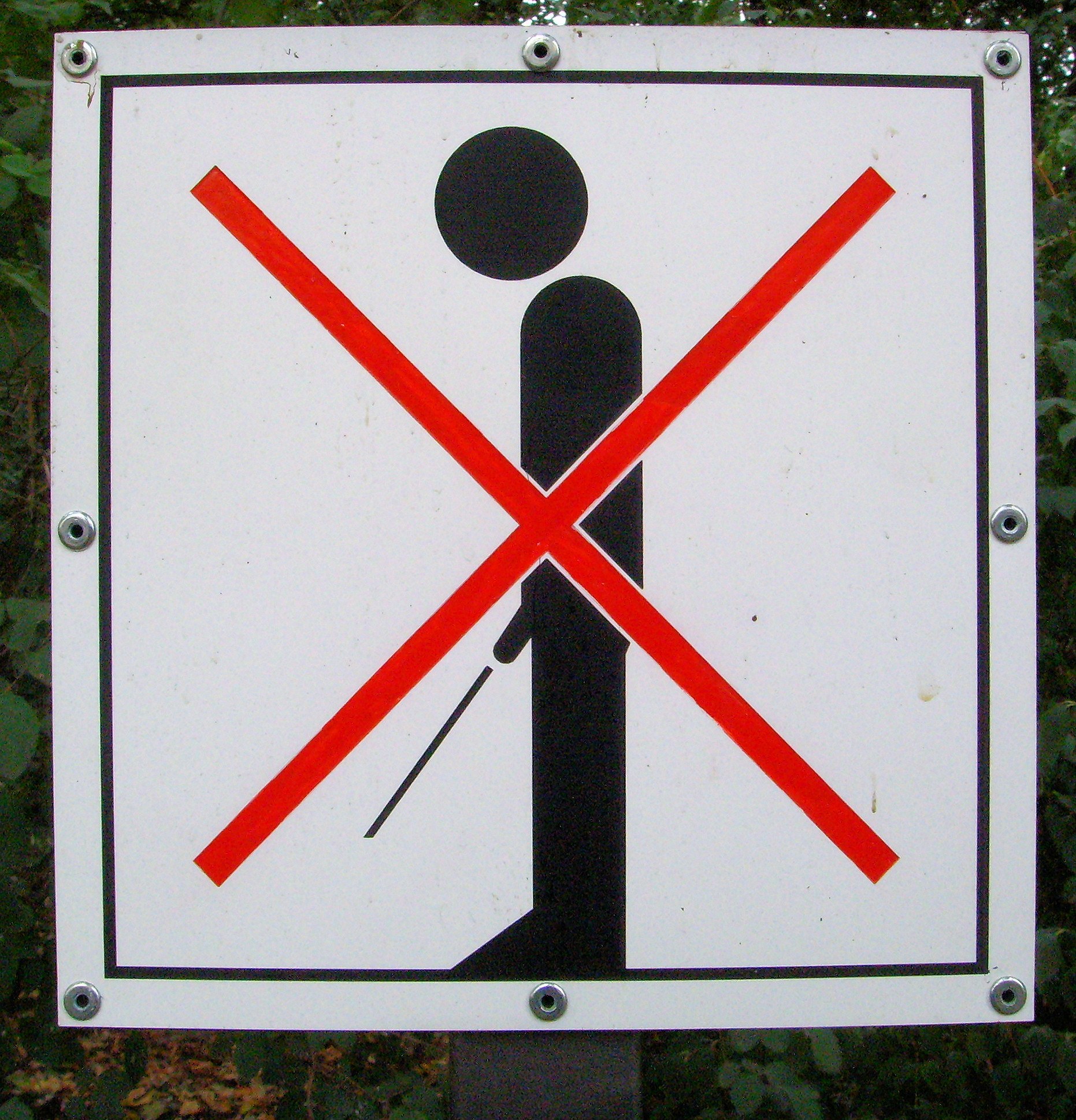 Weißes Parkverbotsschild mit schwarzem Text und rotem Kreuzsymbol, das an einem Pfahl befestigt ist, umgeben von Bäumen in einem bewaldeten Gebiet.