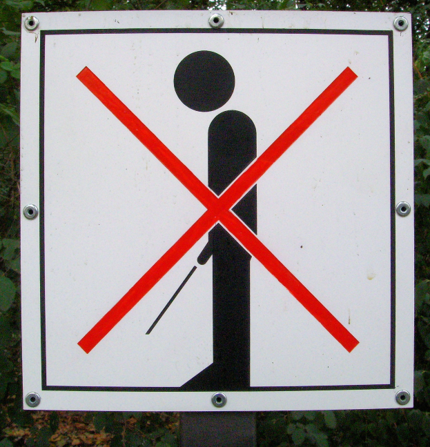 Weißes Parkverbotsschild mit schwarzem Text und rotem Kreuzsymbol, das an einem Pfahl befestigt ist, umgeben von Bäumen in einem bewaldeten Gebiet.