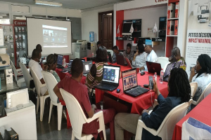 Gruppe von Menschen an einem Tisch mit Laptops und anderen Gegenständen während eines digitalen Marketing-Workshops, mit einem Banner, einem Bildschirm und Deckenleuchten im Hintergrund.