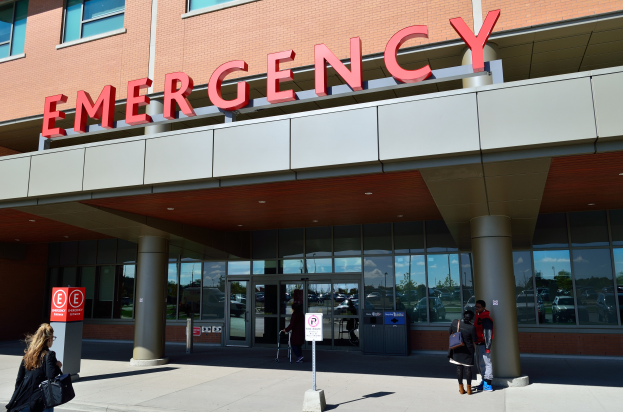 Eine Gruppe von Menschen, die vor einem Glaswand-Notfallgebäude mit Säulen, einem Namensschild, Schildern auf Stangen und einem Mülleimer gehen.