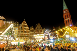 Ein geschäftiger Weihnachtsmarkt in Nürnberg, Deutschland mit Menschen um geschmückte Stände, festliche Lichter, Gebäude mit Fenstern, einen Uhrenturm und einen dunklen Himmel.
