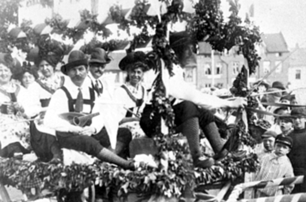 Eine Gruppe von Menschen in traditioneller bayrischer Tracht, einige mit Musikinstrumenten, fährt auf einem mit Laub geschmückten Festwagen in einem Umzug, mit Gebäuden und einem klaren Himmel im Hintergrund, dargestellt in Schwarz-Weiß.