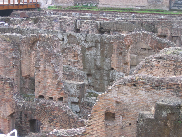 Römerzeitliche Forumruinen mit verwitterten Steinwänden, Bögen und Säulen, grünem Gras, einer hölzernen Brücke mit Geländern auf der linken Seite und fernen Gebäuden im Hintergrund.