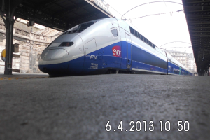 Ein Zug ist in der Mitte des Bildes zu sehen, mit einem Bahnsteig und Text unten, Gebäuden und einem Dach links und Gebäuden, Dach, Kabeln und Himmel oben.