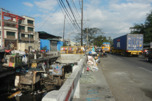 Eine stadtstraßenfüllende Müllhalde mit Fahrzeugen, Gebäuden, Bäumen, Polen, Drähten, unter einem bewölkten Himmel.