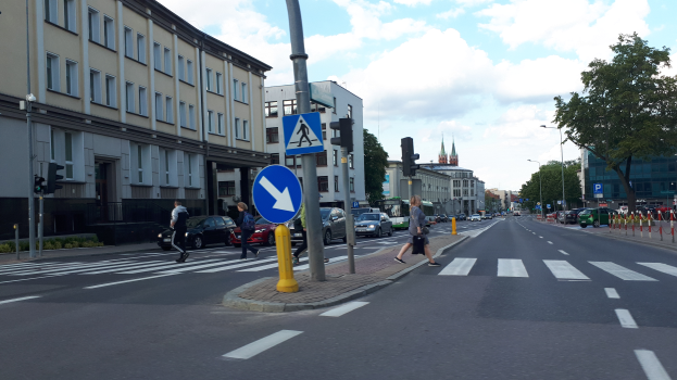 Eine Stadtstraßenszene mit Menschen, die an einer Ampel über die Straße gehen, Fahrzeugen auf der Straße, Verkehrszeichen, Lichtmasten, Bäumen, Gebäuden mit Fenstern und einem bewölkten Himmel im Hintergrund.