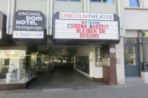 Das Lincoln Theater in Berlin, Deutschland, ein Gebäude mit Glasfenstern und -türen und einer Tafel mit Text darauf, mit verschiedenen Objekten im Inneren, die einen belebten Stadtbild Eindruck vermitteln.