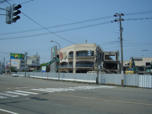 Eine Stra√e in einer Stadt mit fahrenden Autos, Strommästen, Laternen und einer Ampel auf der linken Seite, Gebäude, Bäume und Himmel im Hintergrund und ein abbruchreifes Gebäude im Vordergrund.