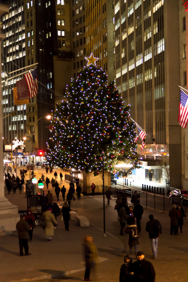 Ein Weihnachtsbaum, geschmückt mit Menschen, steht in der Mitte einer Straße, flankiert von zwei Fahnenmasten, mit beleuchteten Gebäuden im Hintergrund.