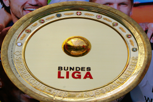 Ein Mann mit einem goldenen Schild mit der Aufschrift "Bundesliga", mit einem Banner im Hintergrund mit drei Personen.