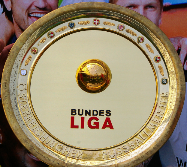 Ein Mann mit einem goldenen Schild mit der Aufschrift "Bundesliga", mit einem Banner im Hintergrund mit drei Personen.