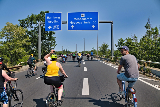 Eine Gruppe von Radfahrern mit Helmen fährt auf einer Straße mit einer Begrenzung auf einer Seite und Bäumen auf der anderen, unter einem klaren blauen Himmel mit Laternen im Hintergrund; ein Schild oben könnte eine Radtour in Hamburg anzeigen.