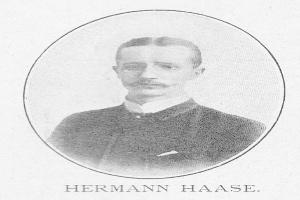 Schwarz-weißes Porträt des deutschen Komponisten und Pianisten Hermann Haase in einem Anzug, mit Schnurrbart und ernstem Gesichtsausdruck, mit deutschem Text unten.