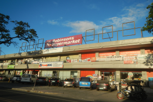 Ein Robinsons Supermarkt in Singapur mit Autos auf der Straße, einer Person auf dem Gehweg, einem Fahrrad, Pfosten, einem Schild, einem Metallrahmen, Bäumen und einem bewölkten Himmel.
